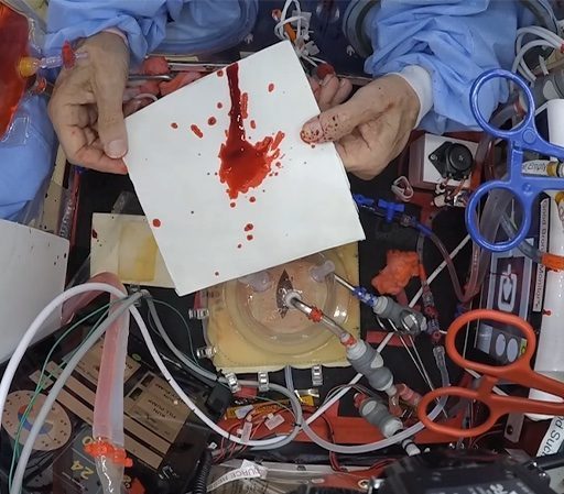 A cluttered workspace with electronic components and a hand holding a card splattered with red liquid.