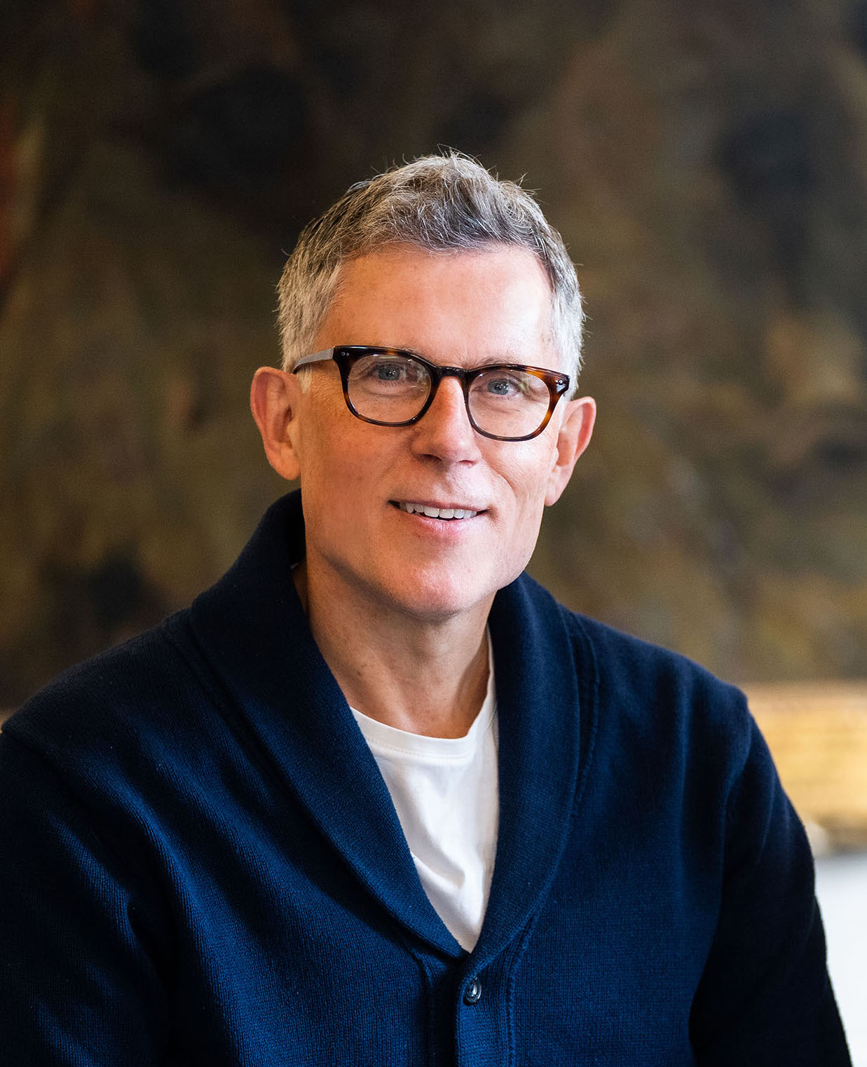 Middle-aged caucasian man with glasses, wearing a navy cardigan and white shirt, smiling and sitting in front of an abstract painting.