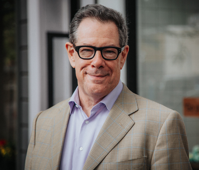 Middle-aged man smiling, wearing glasses, a beige blazer, and a purple shirt, standing outside a building.