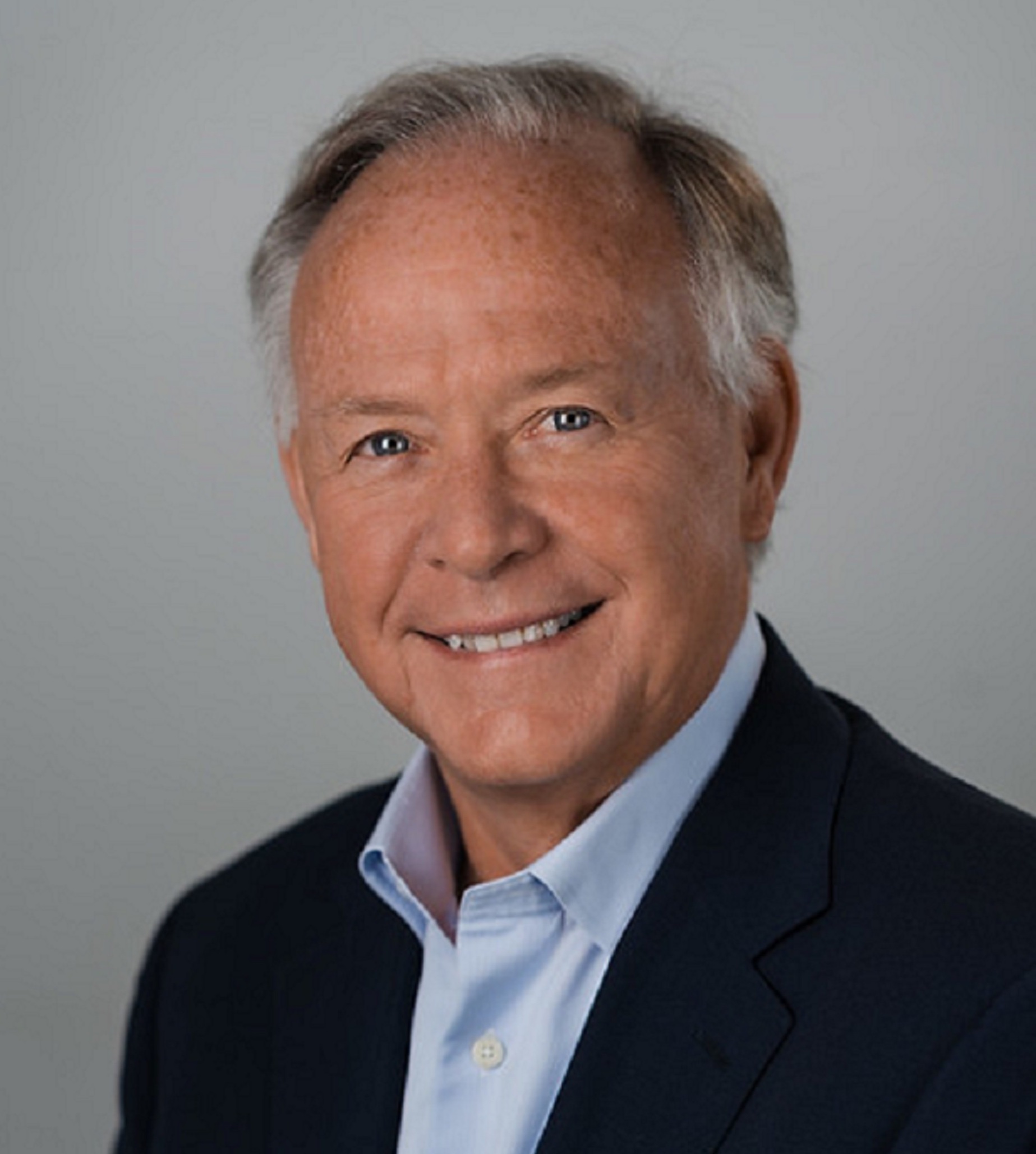 Professional headshot of an elderly man with fair skin smiling, wearing a dark suit jacket and a light blue shirt against a gray background.
