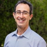 A smiling man with glasses wearing a blue patterned shirt, standing outdoors with green foliage in the background.