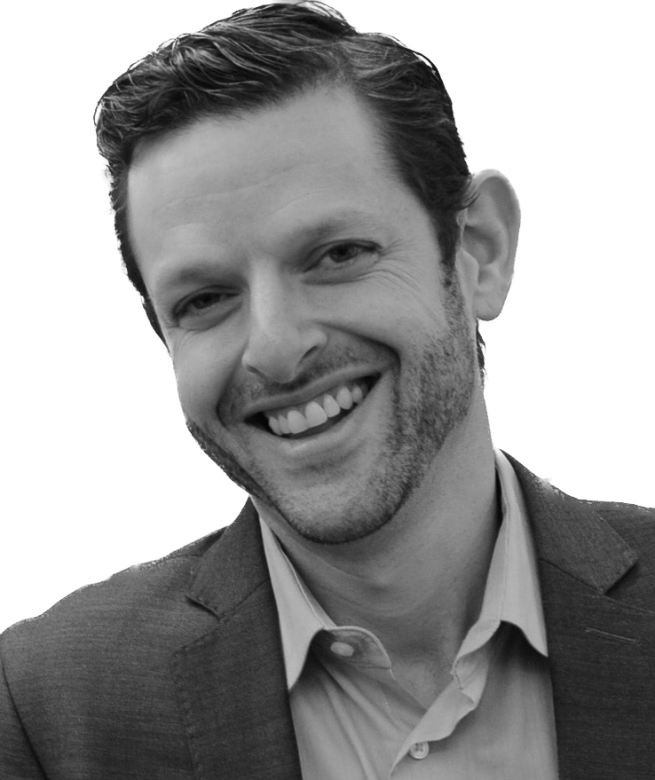 A black and white image of a smiling man with stubble, wearing a suit jacket and a collared shirt.