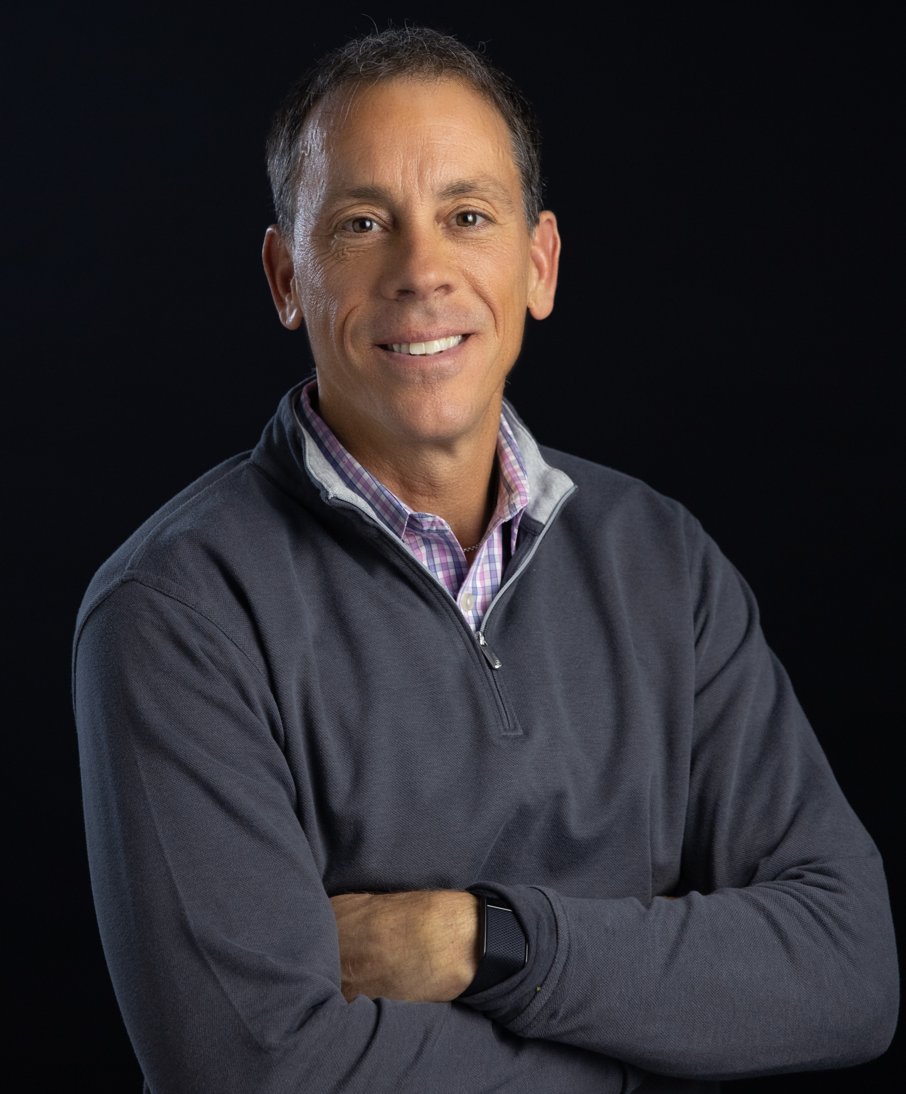 A middle-aged man smiling, wearing a grey pullover and a checkered shirt, arms crossed, against a black background.