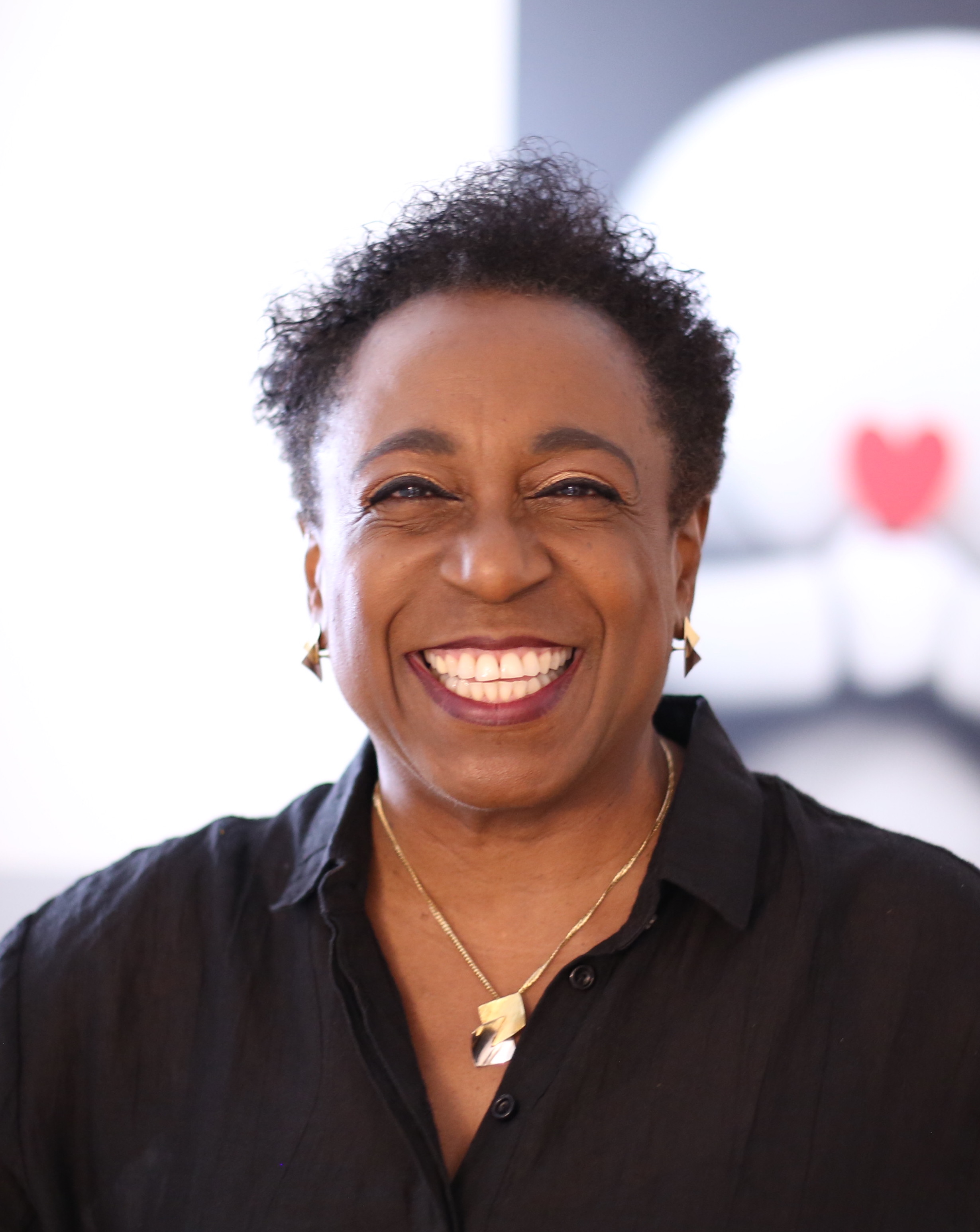 A joyful middle-aged woman with short curly hair, wearing a black shirt and earrings, smiles warmly at the camera.