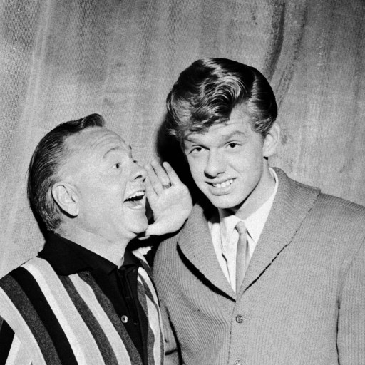 Black and white photo of an older man in a striped jacket whispering to a grinning young man in a suit.