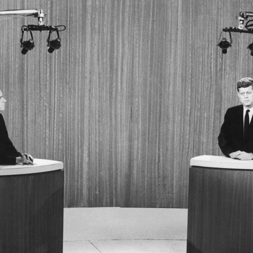 Two men from different political parties in suits stand at podiums under studio lights, participating in a televised debate.
