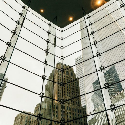 View of a cityscape through a large glass window framed by steel beams, showcasing high-rise buildings burdened with debt under an overcast sky.