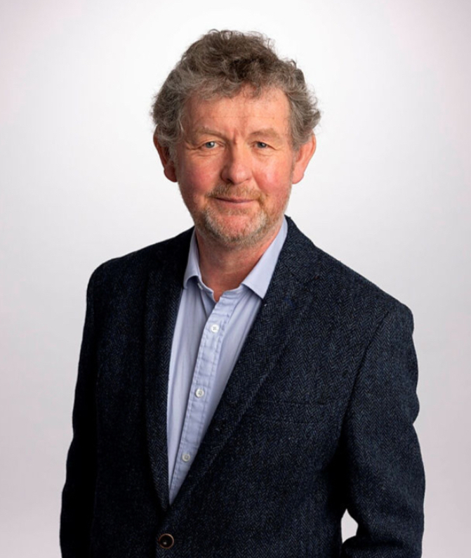 An older man with curly gray hair, smiling slightly, wearing a blue blazer and gray shirt, standing against a light background.