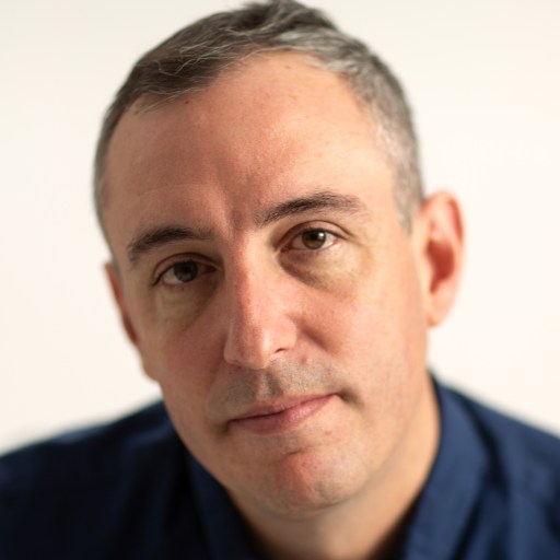 A man with short gray hair and a serious expression is looking directly at the camera, wearing a blue shirt against a plain background.