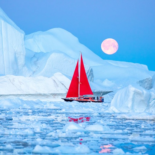 A sailboat with a red sail navigates through icy waters surrounded by large icebergs under a full moon.