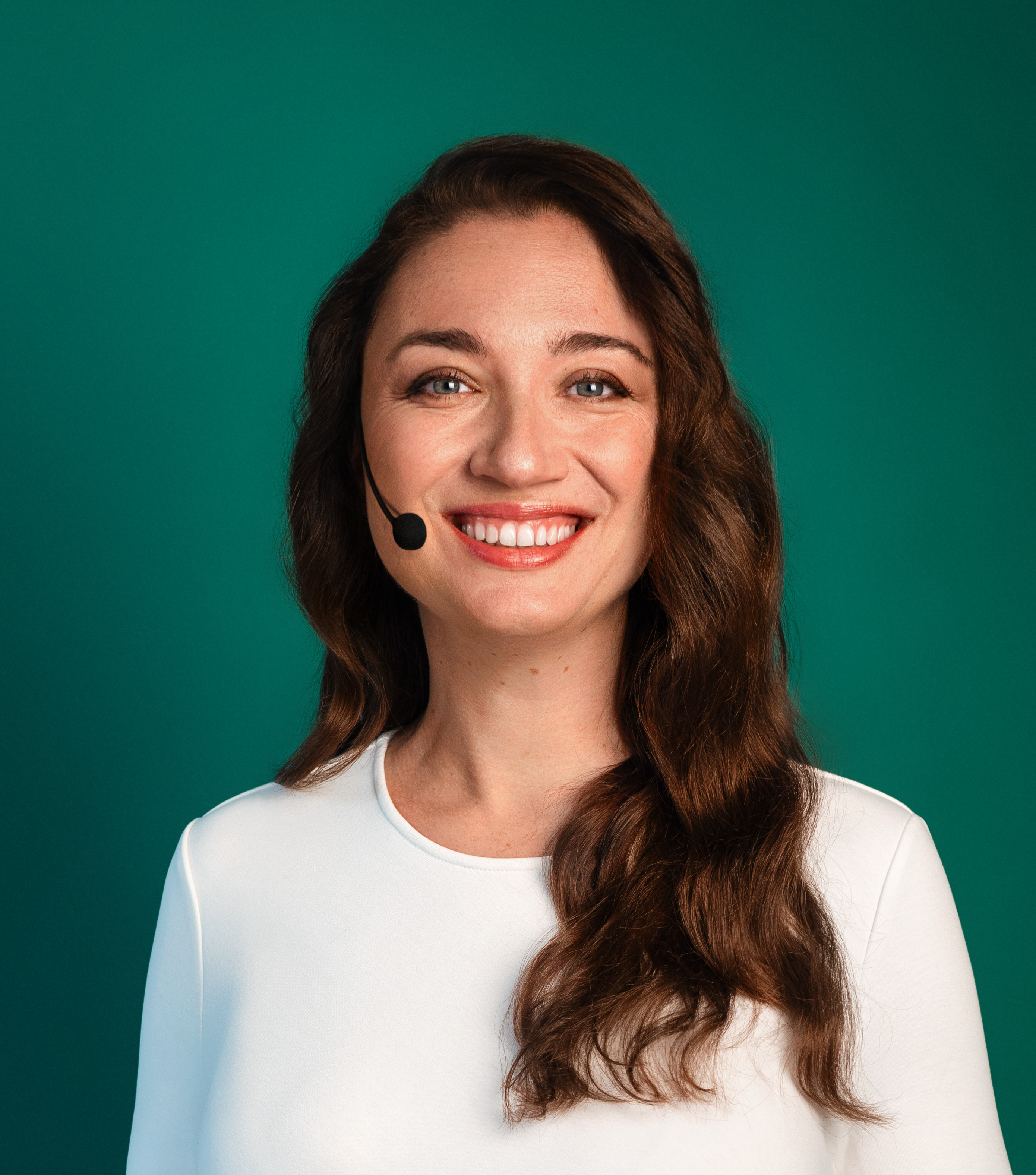 A woman with long brown hair, wearing a white top and a headset, smiles at the camera against a green background.