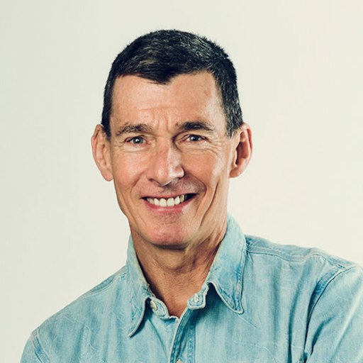 A middle-aged man with short dark hair, wearing a light blue denim AI shirt, smiling at the camera against a plain light background.