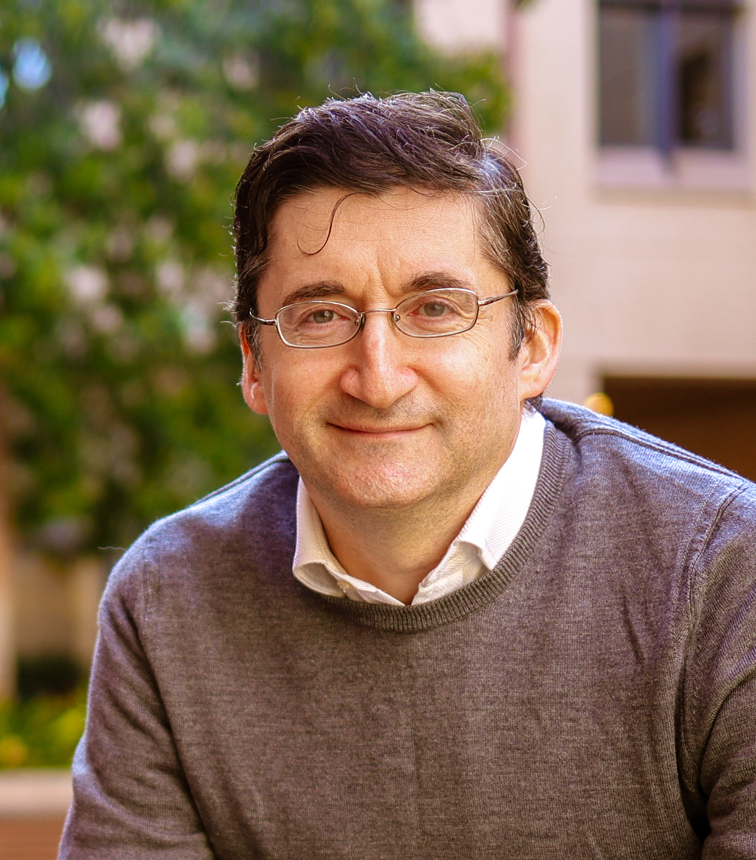 A man with glasses, short dark hair, and a gray sweater over a white collared shirt smiles outdoors in front of a blurred background of greenery and a building.