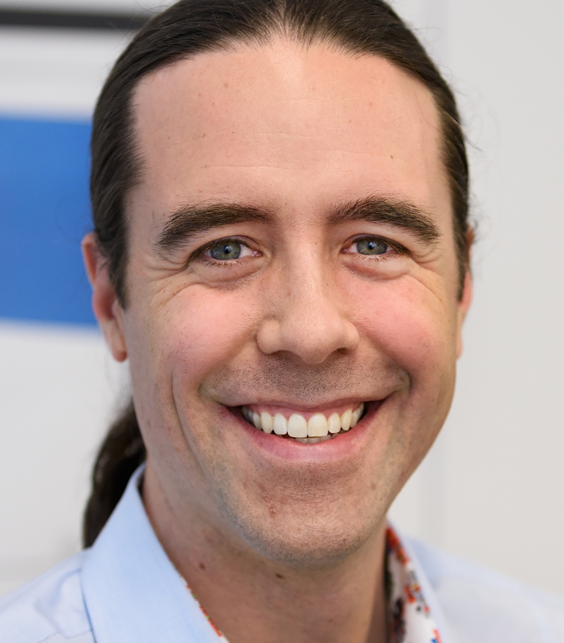 A smiling person with long dark hair tied back, wearing a light blue shirt, in a close-up shot.