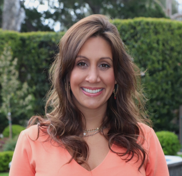 A woman with long brown hair smiling, wearing an orange top and standing outdoors in front of green foliage.