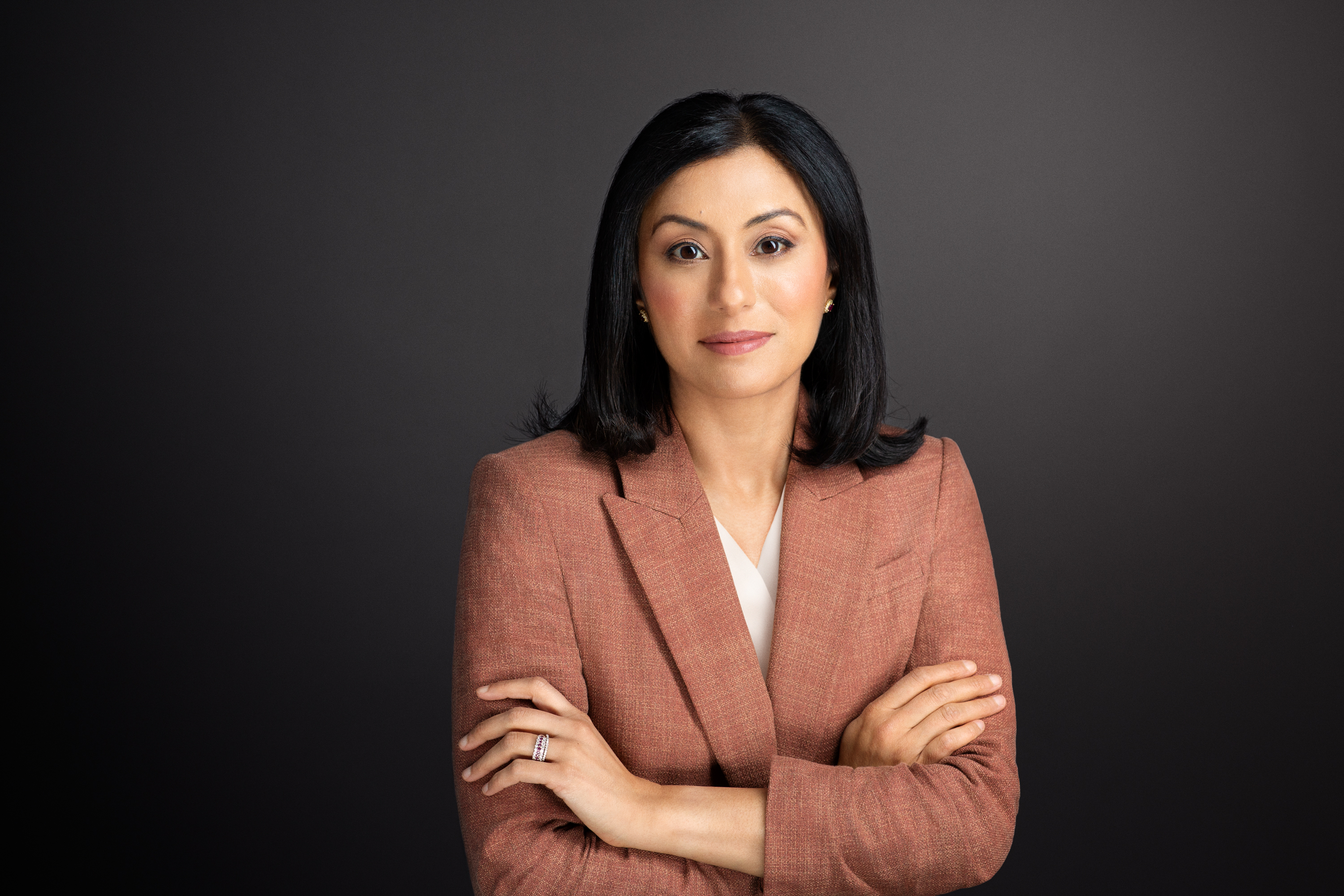 A woman in a rust-colored blazer stands with her arms crossed against a dark gray background, looking directly at the camera.