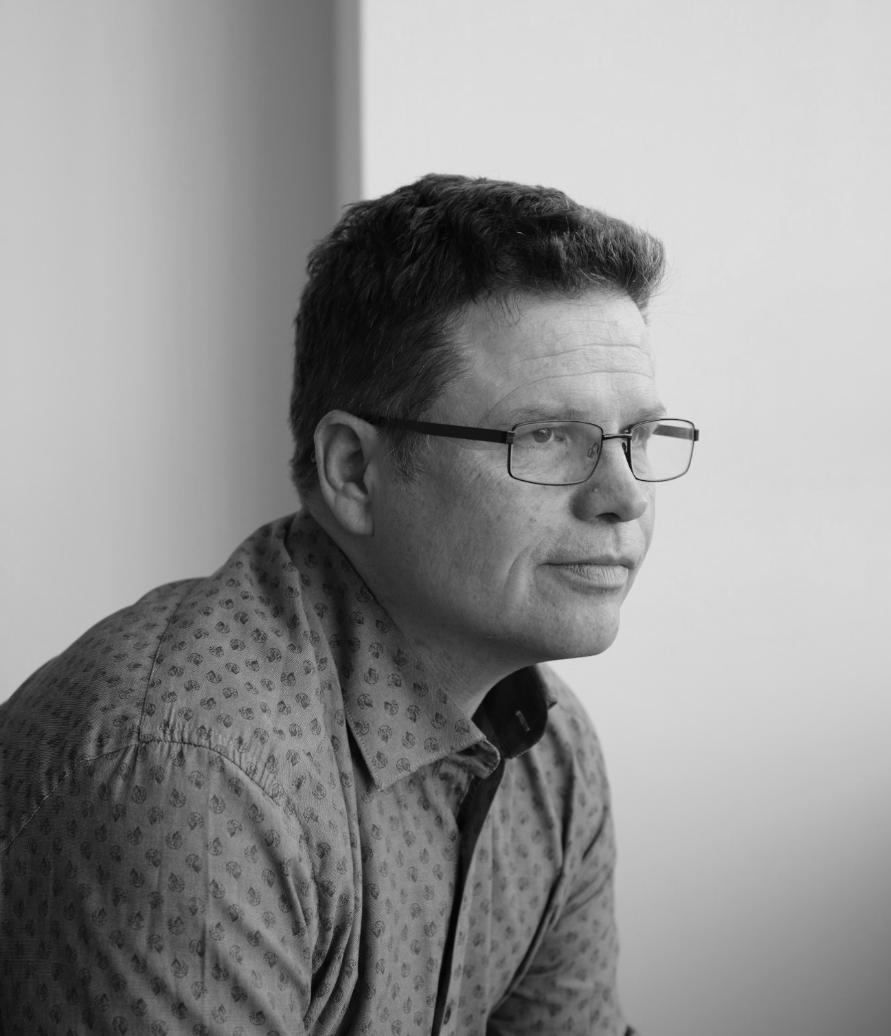 A man wearing glasses and a patterned shirt looks pensively to the side in a black and white photo.
