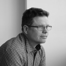 A man wearing glasses and a patterned shirt looks pensively to the side in a black and white photo.
