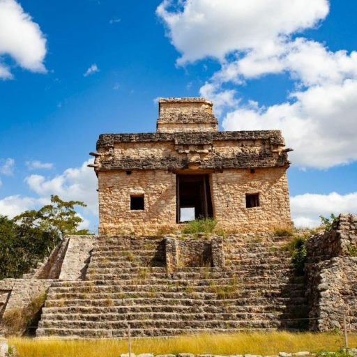 Ancient stone structure with stairs under a blue sky with scattered clouds, surrounded by vegetation.