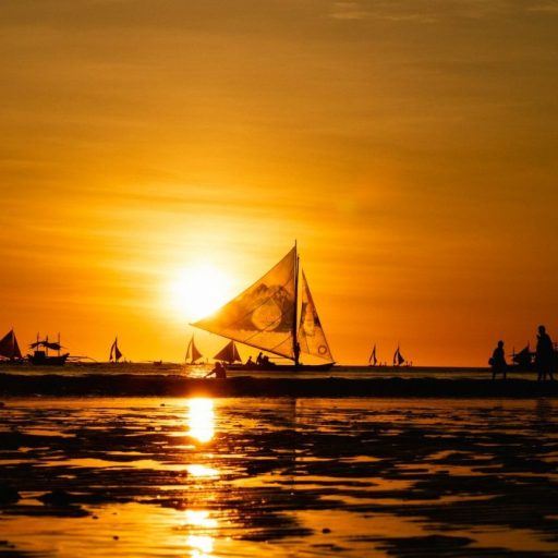 A sailboat is silhouetted against a vibrant sunset over a calm sea, with other boats and their reflections visible in the background. The sky and water have a golden hue.