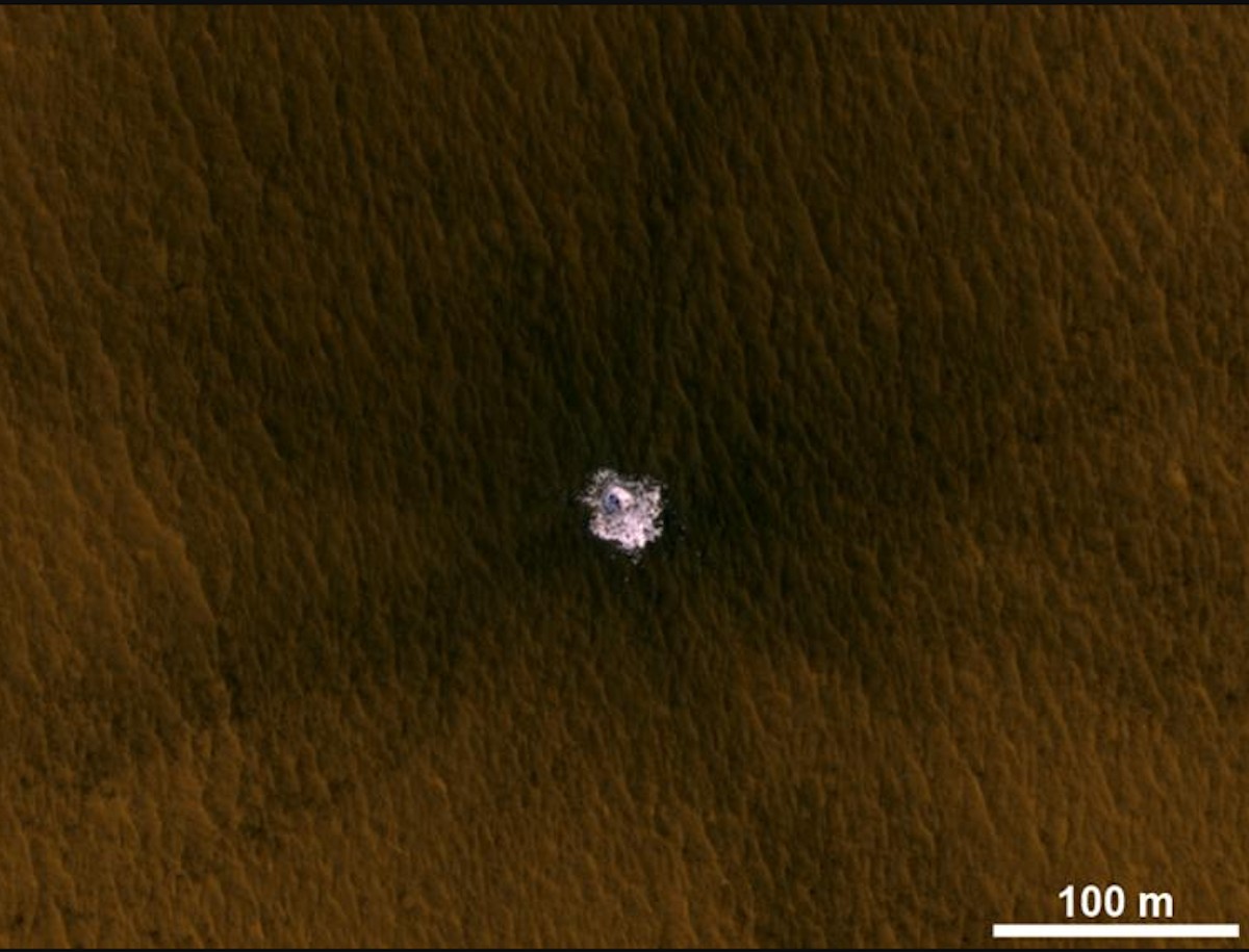 Aerial view of an impact crater on a brown landscape, showcasing a central white area where the impact occurred. A scale bar indicating 100 meters is present in the bottom right corner.