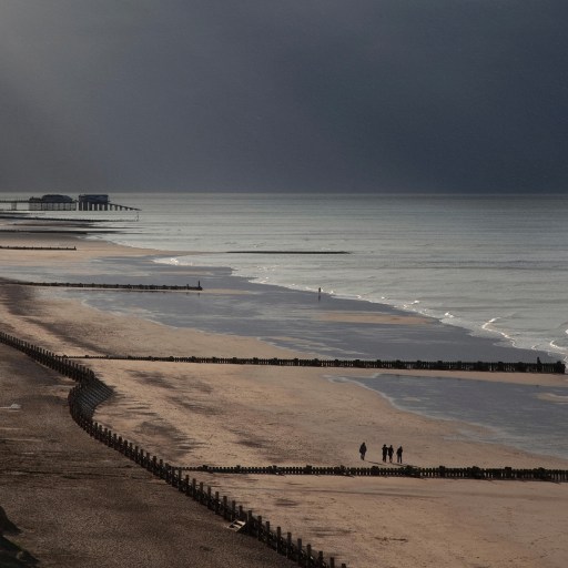 A serene beach scene at dusk with a distant pier, a few people walking along the shoreline, and gentle waves hitting the sand. The sky is cloudy with rays of light breaking through, hinting at the subtle potential for tidal power beneath the surface.