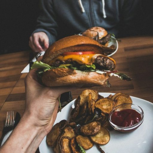 A person holding a burger with fries and ketchup on a wooden table.