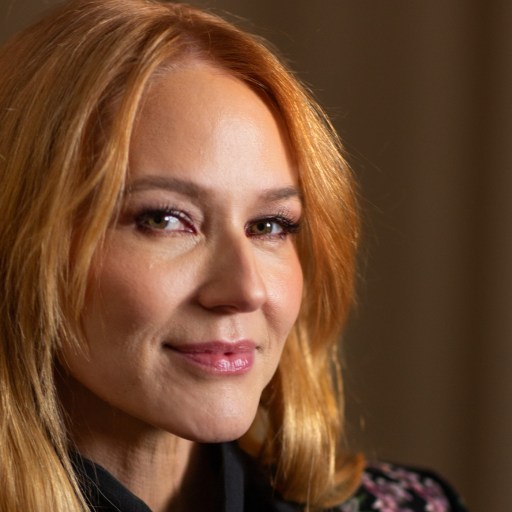 A person with long, light brown hair is looking at the camera with a soft smile, wearing a dark colored outfit.