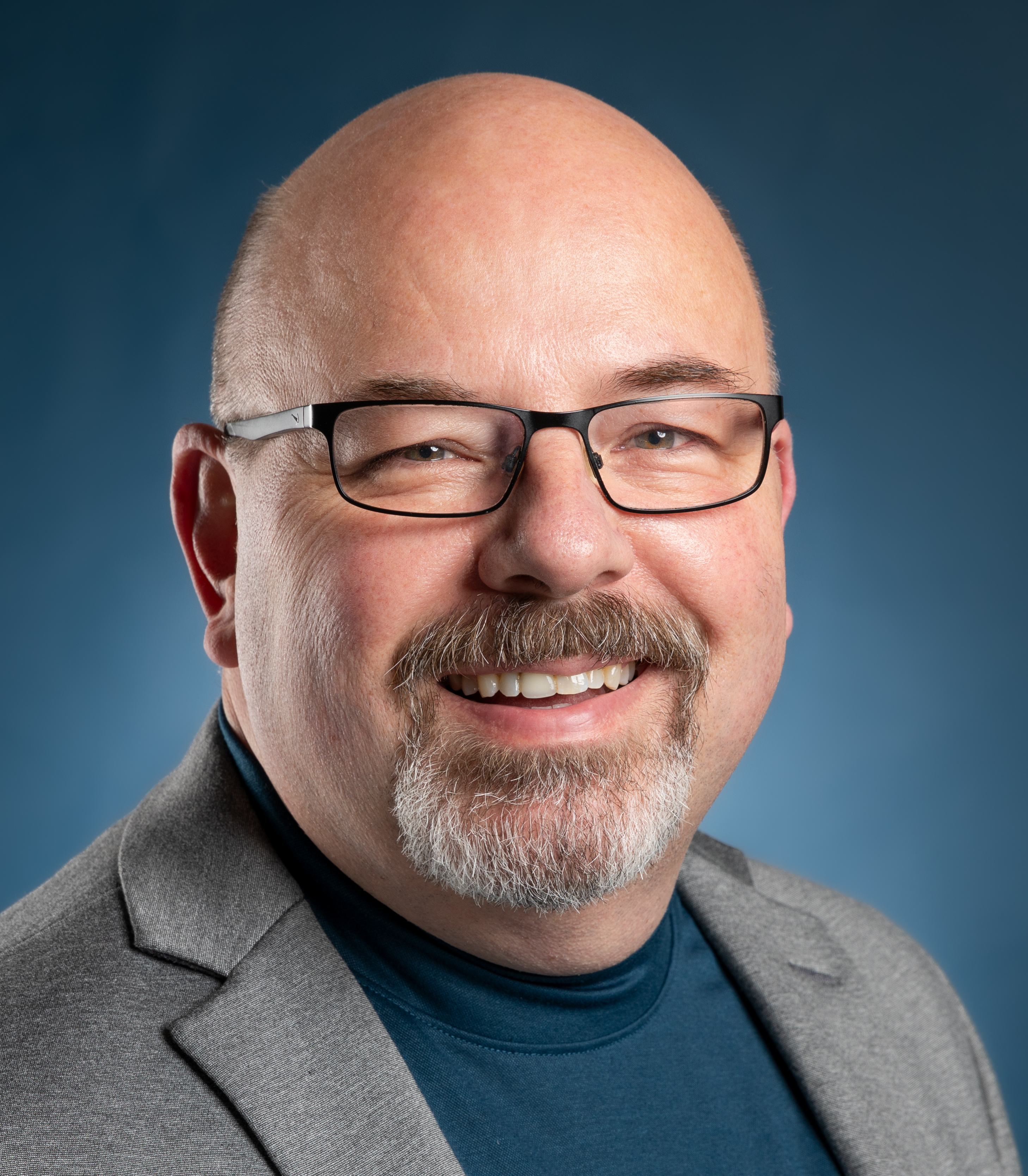 Bald man with glasses, a goatee, and a mustache smiles while wearing a gray blazer and a teal shirt against a blue background.