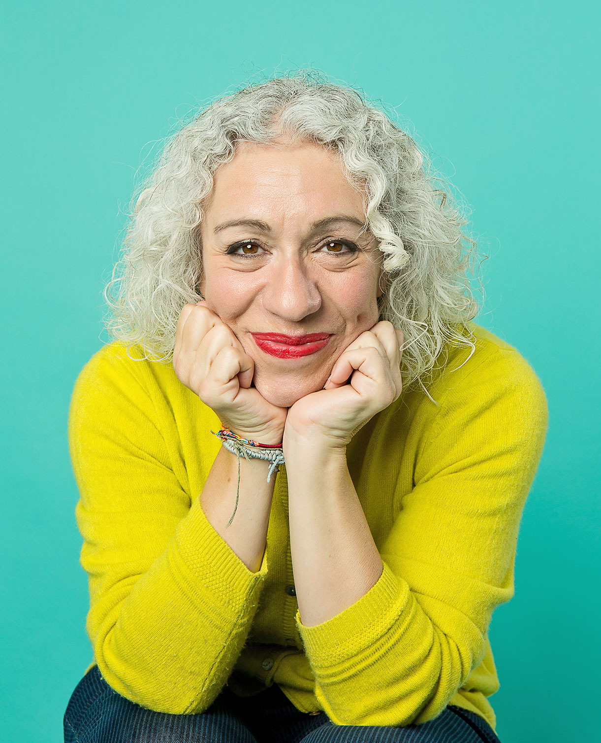 A person with curly gray hair and wearing a yellow sweater rests their head on their hands, smiling against a teal background.