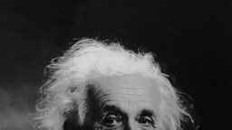 Black and white close-up photograph showing the upper part of an elderly person’s face and their white, unruly hair against a dark background.