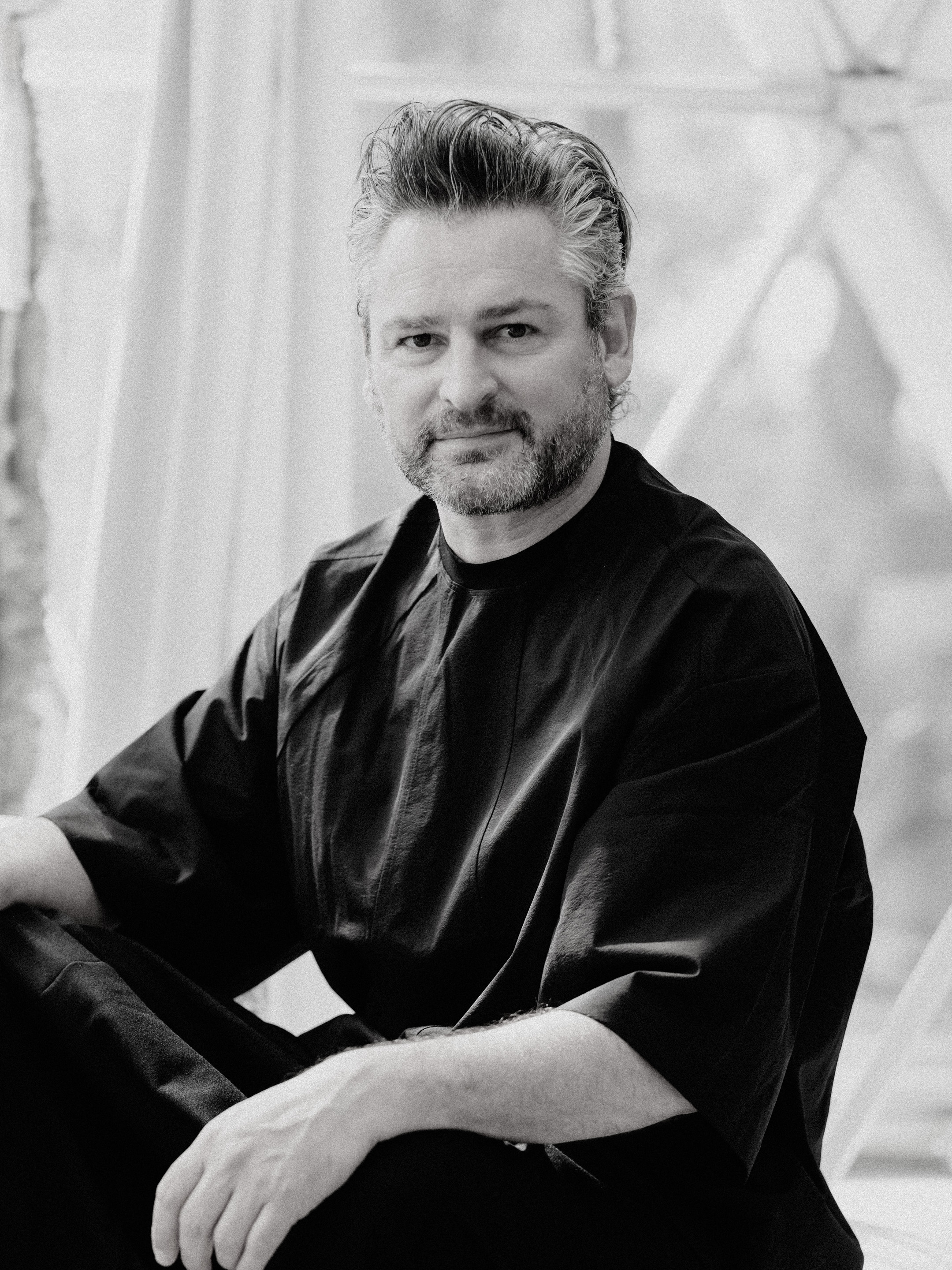 A man with short, graying hair and a beard sits indoors wearing a loose, dark shirt. The background is softly lit and blurred.