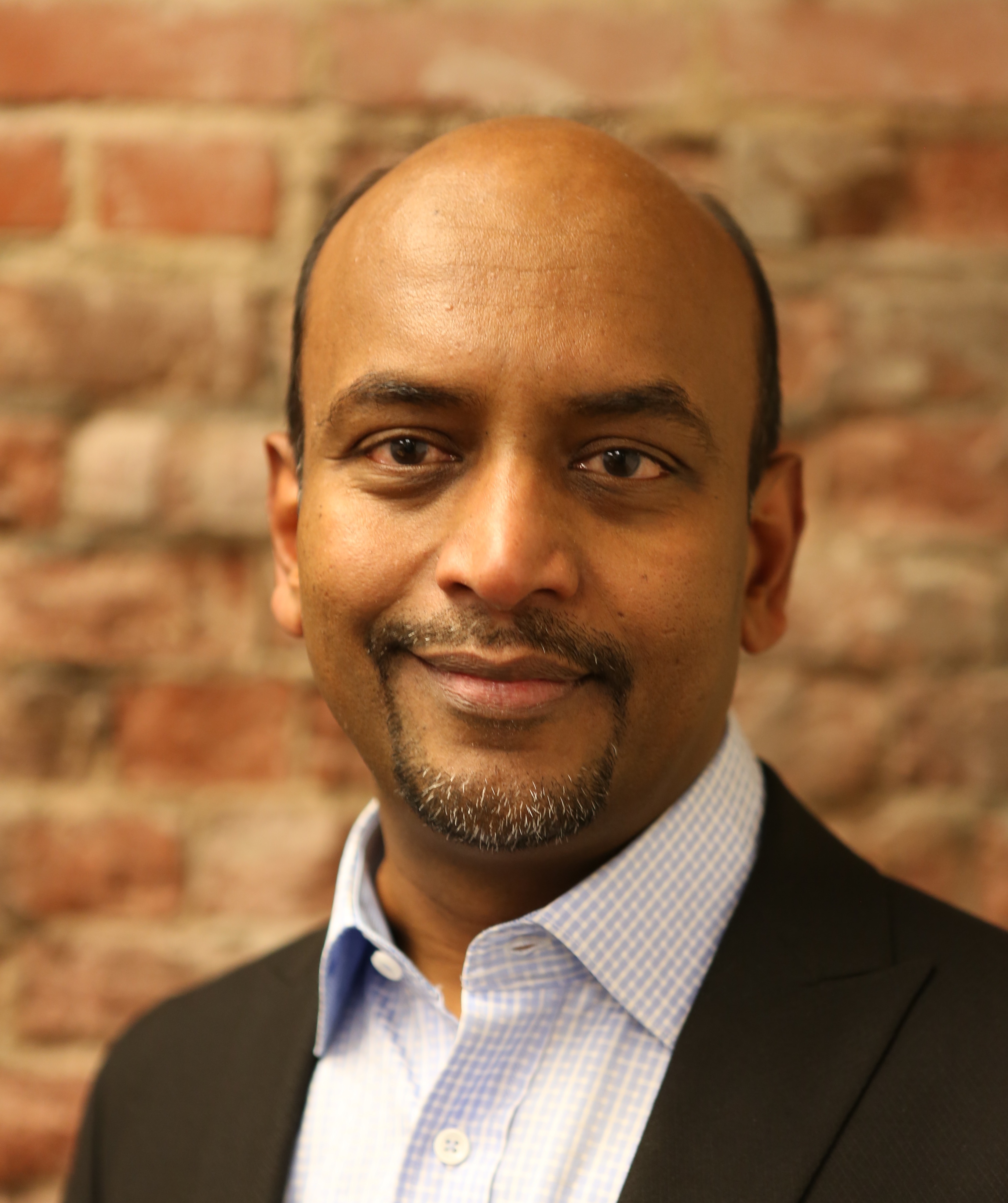 A man with a shaved head, wearing a suit jacket and blue shirt, stands in front of a brick wall, smiling slightly.