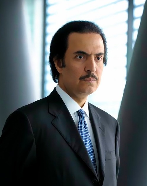 A man in a suit and tie stands indoors against a backdrop of vertical blinds.