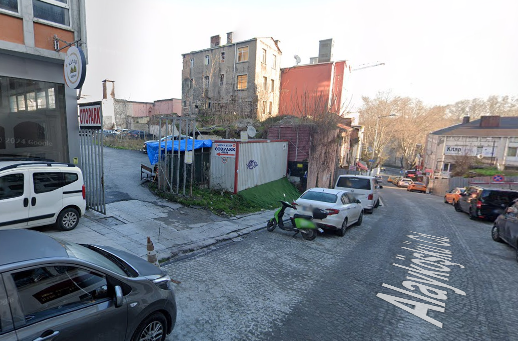 A cobblestone street with parked vehicles, a small gated lot with a tent, and surrounding buildings, taken near the junction of Alayk&ouml;şk&uuml; Cd.