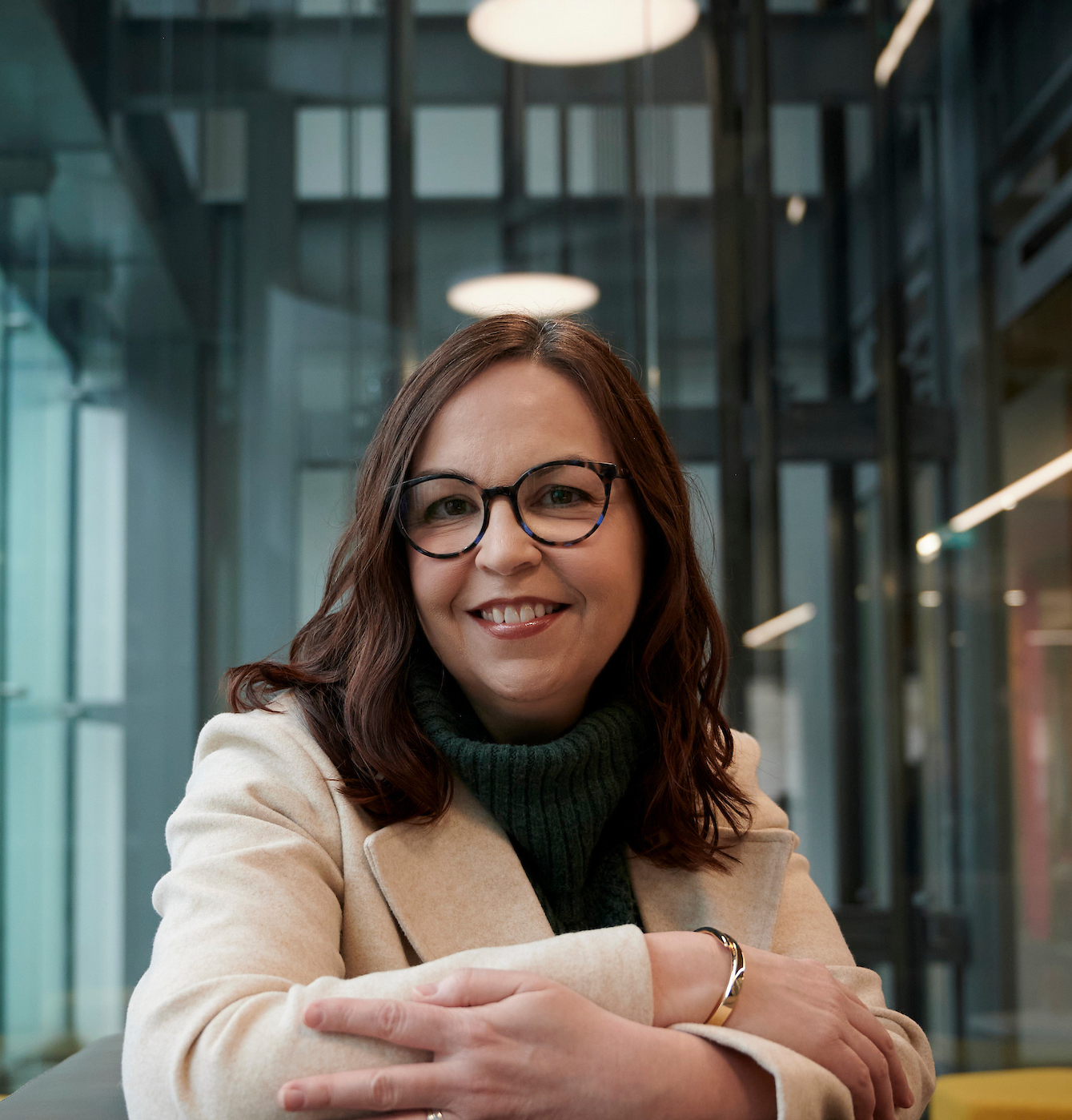 A woman with long brown hair and glasses, wearing a light coat and green turtleneck, smiles while seated indoors with modern, glass-walled surroundings.