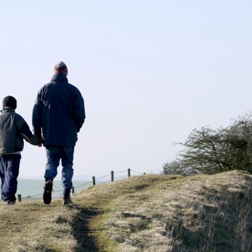 Two people walk hand in hand on a grassy hilltop trail, wearing winter jackets and hats. Sparse trees and a distant landscape are visible under a clear sky.