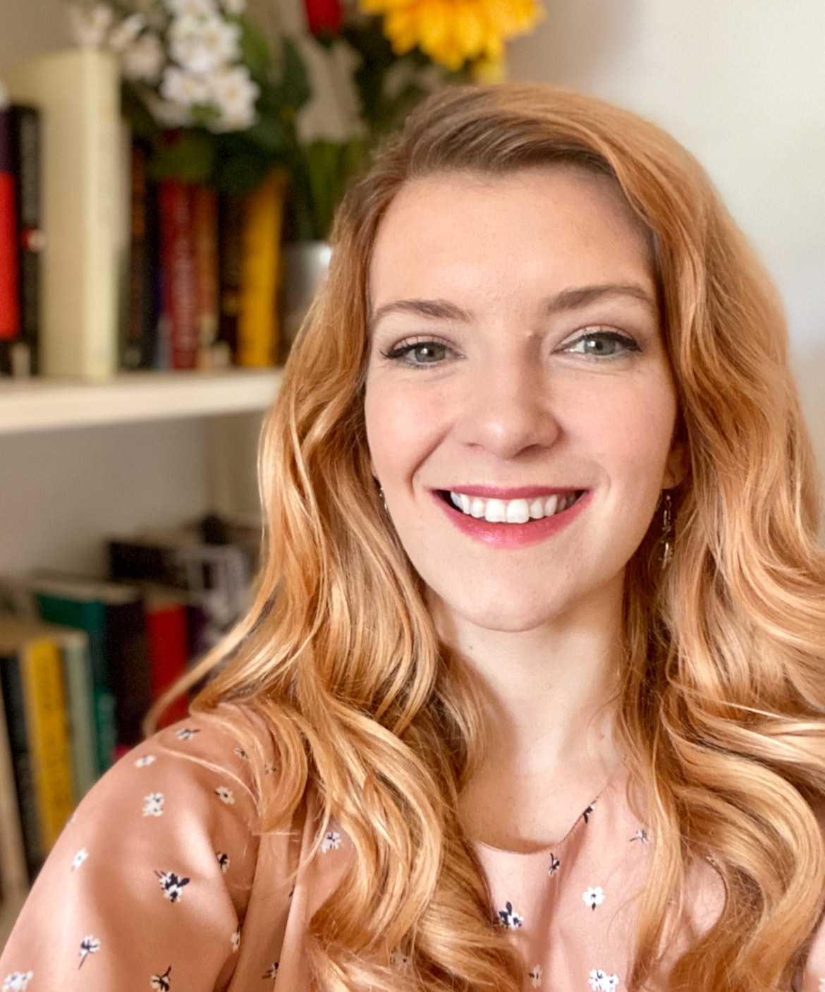 A woman with long wavy hair smiles while posing in front of a bookshelf with colorful books and a vase of flowers.