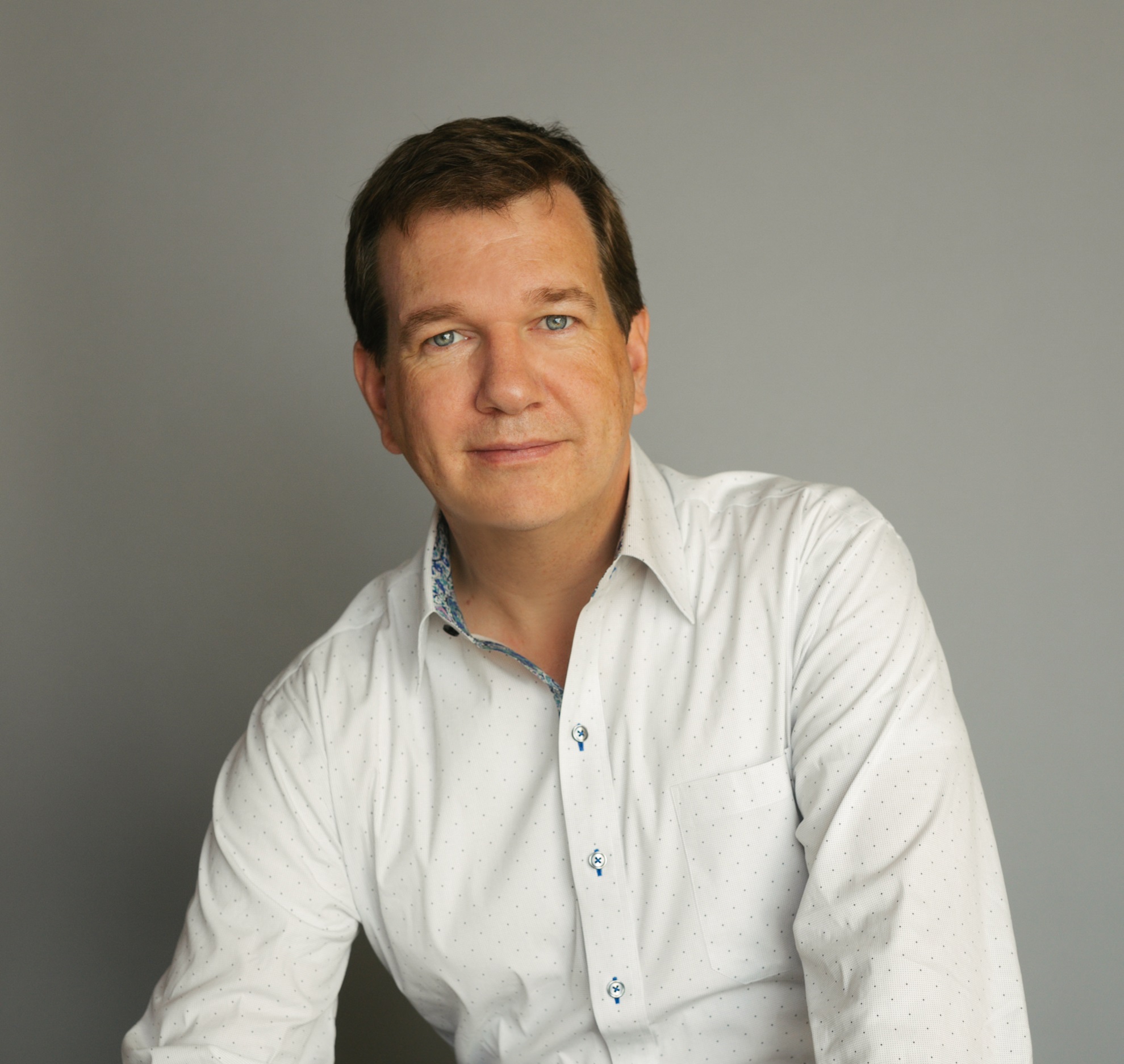 A man with short brown hair and blue eyes wearing a white button-up shirt sits against a gray background.