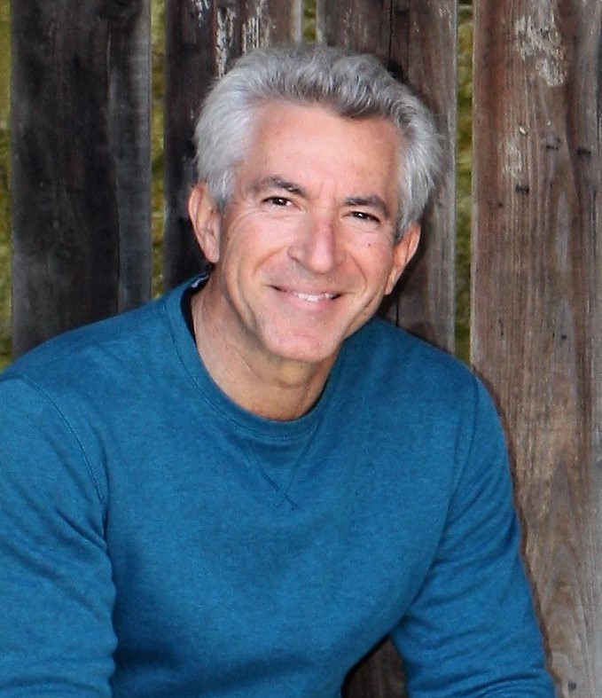 A man with gray hair wearing a blue sweater smiles while sitting in front of a wooden fence.