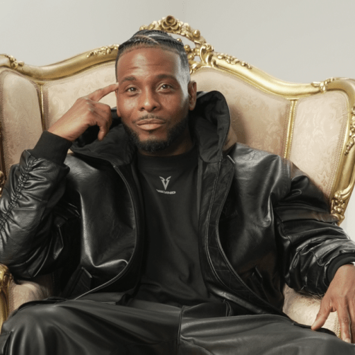 A man in a black leather outfit sits on an ornate, gold-trimmed chair, with one hand touching his temple and the other resting on the armrest.