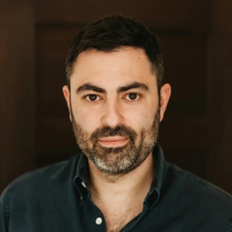 A man with short dark hair and a beard, wearing a dark button-up shirt, looks directly at the camera against a dark blurred background.