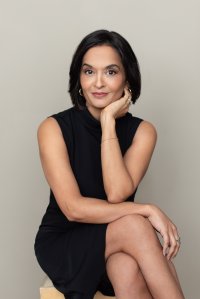 A woman with short black hair is sitting, wearing a black sleeveless dress. She has her chin resting on her hand and she is smiling slightly, with a neutral background.