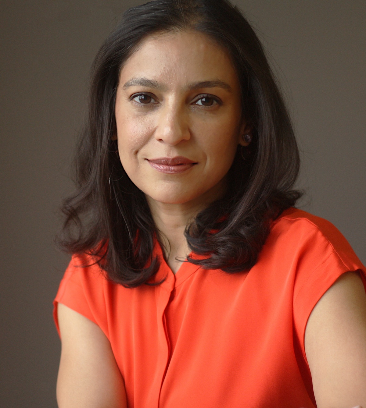 A woman with shoulder-length dark hair wearing an orange blouse poses for a photo against a plain background.