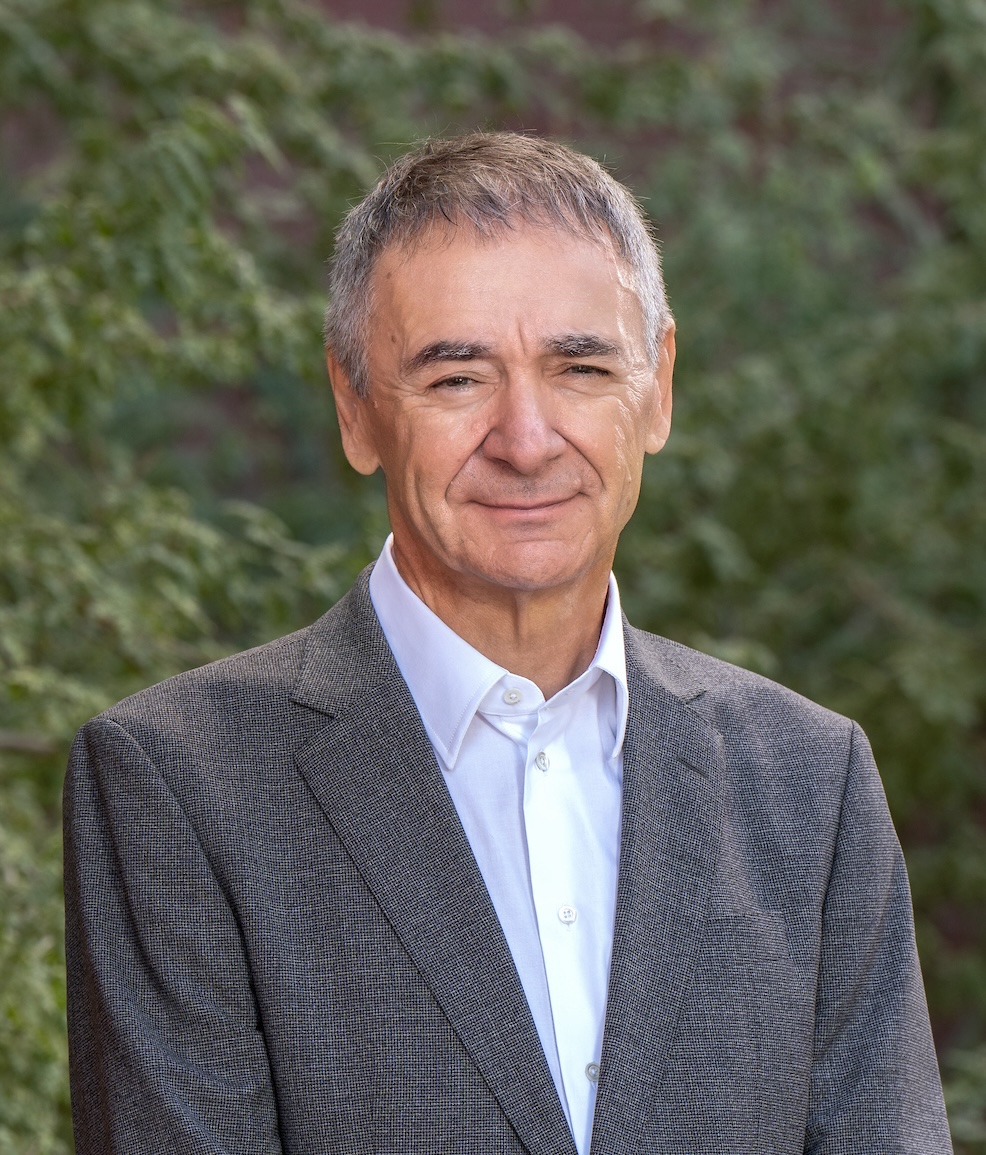 A man with short gray hair wearing a gray suit and white shirt stands outdoors, with green foliage in the background.