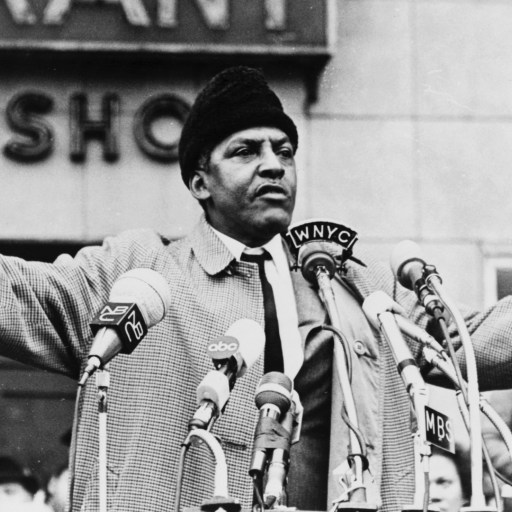A man standing behind multiple microphones gestures while speaking to a crowd in an outdoor setting, with a group of people and a building in the background.