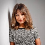 Person with shoulder-length brown hair and bangs, wearing a short-sleeve gray checkered dress, standing against a light gray wall.