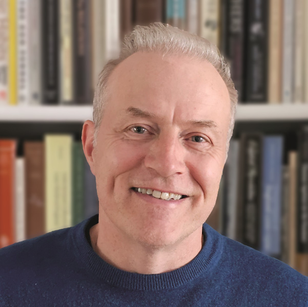 A man with short, light-colored hair and a blue shirt is smiling in front of a bookshelf filled with assorted books.