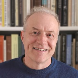 A man with short, light-colored hair and a blue shirt is smiling in front of a bookshelf filled with assorted books.