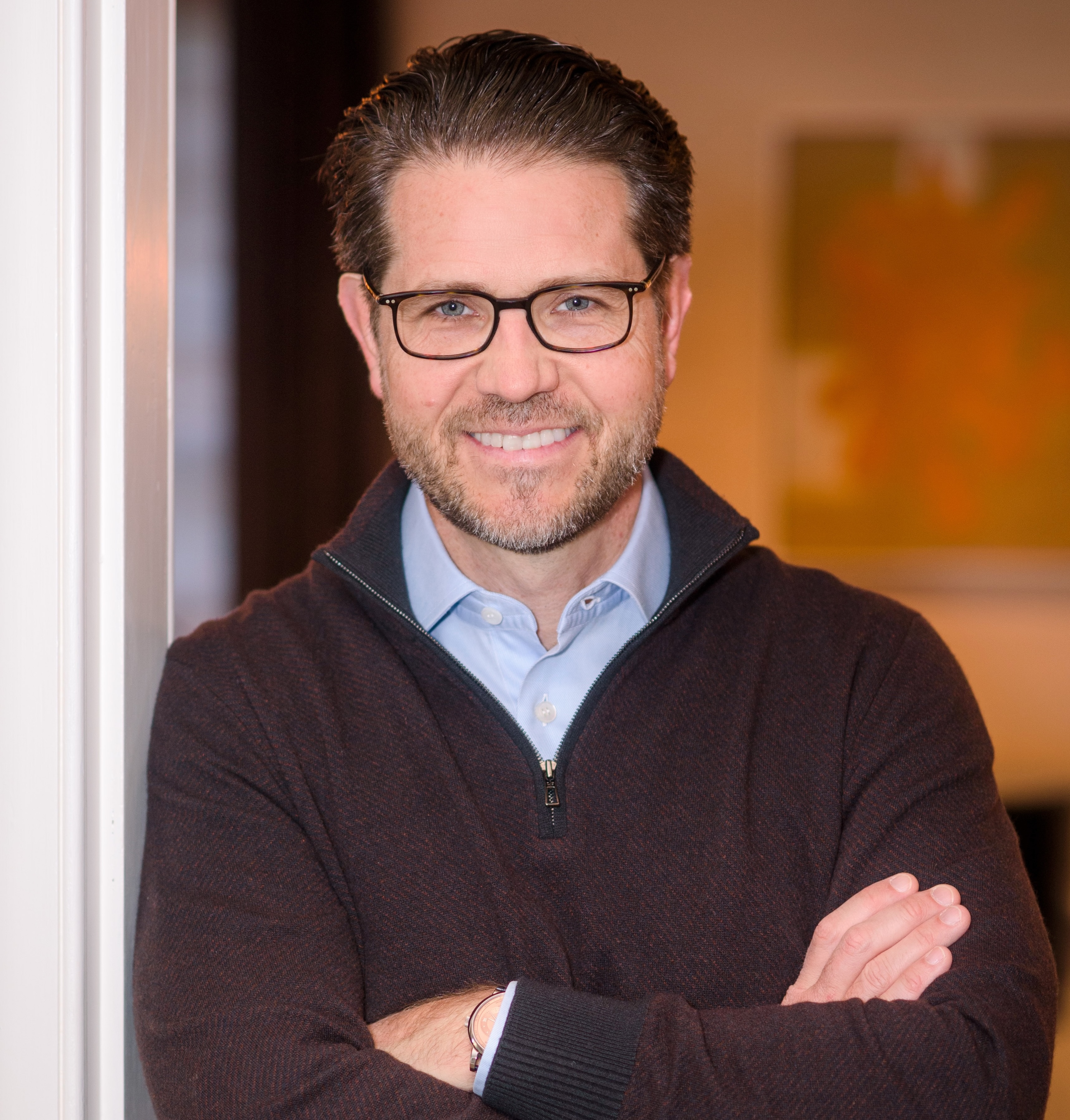 A man with glasses and a beard stands indoors with his arms crossed, wearing a blue collared shirt and a dark sweater.